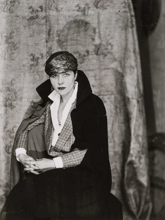 A black-and-white portrait of an elaborately dressed White woman seated in a chair with a coat draped over her shoulders.