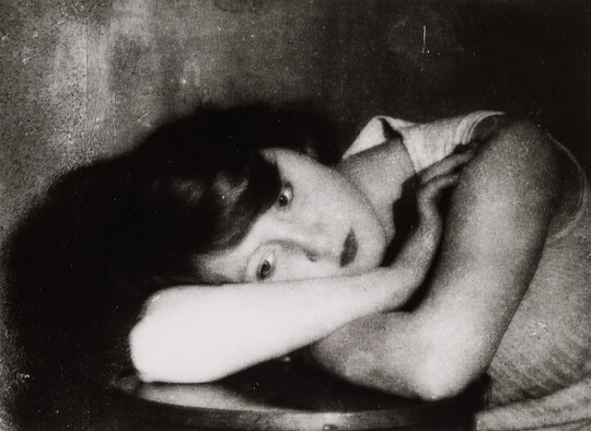 A black-and-white portrait photograph of a White woman leaning over a table and resting her head on her arms.