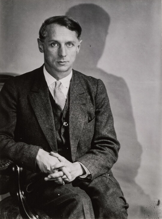 A black-and-white portrait photograph of a White man in a suit sitting in a chair and gazing directly at the camera with his hands folded in his lap.
