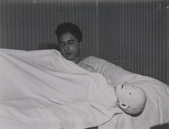 A black-and-white photograph of a White man lying under a white sheet next to a mannequin head.