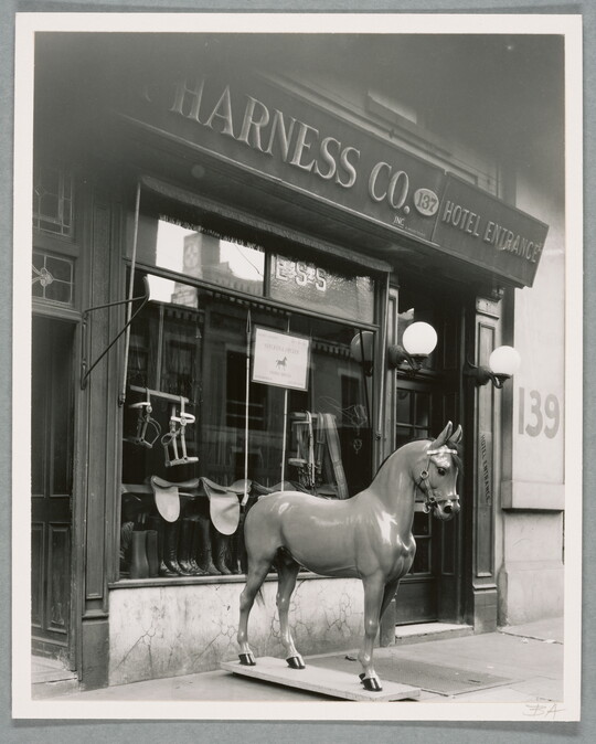 A black-and-white photograph of a storefront with a horse statue on the sidewalk in front of a display window.