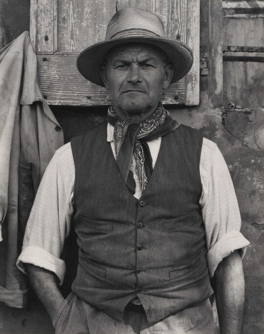A black-and-white portrait photograph of a medium-skinned man from the waist up wearing a hat, bandanna tied around his neck, vest, and a shirt rolled to his elbows.