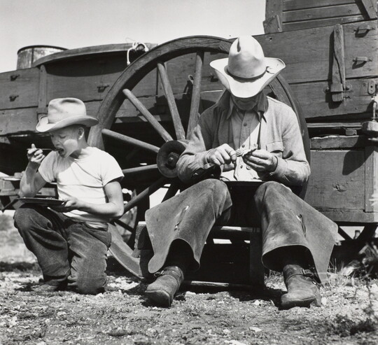 A black-and-white photograph of a White man and boy in cowboy hats sitting on the ground eating next to a wagon. 