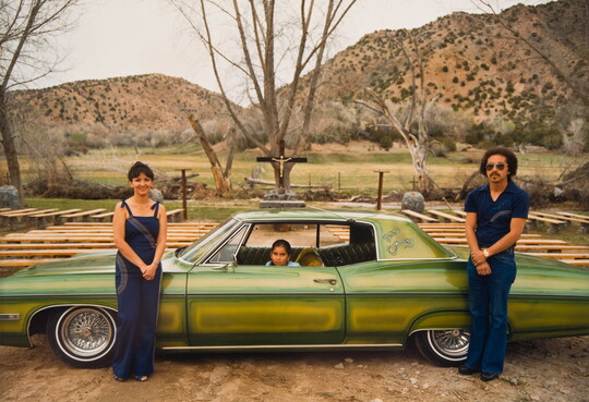 A color photograph of two adults standing in front of a green car that's in profile with a child sitting in the driver's seat.