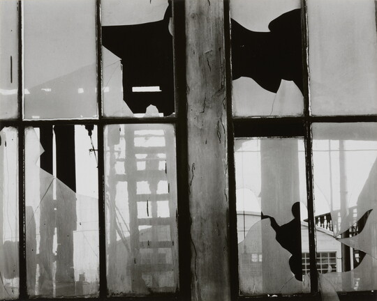 A black-and-white photograph of broken window panes in what looks like a factory building.