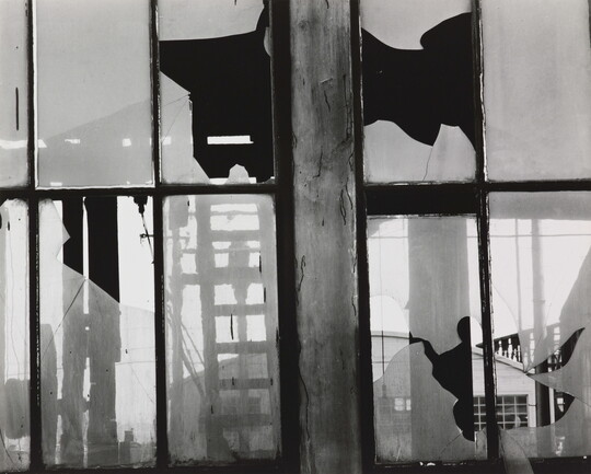 A black-and-white photograph of broken window panes in what looks like a factory building.