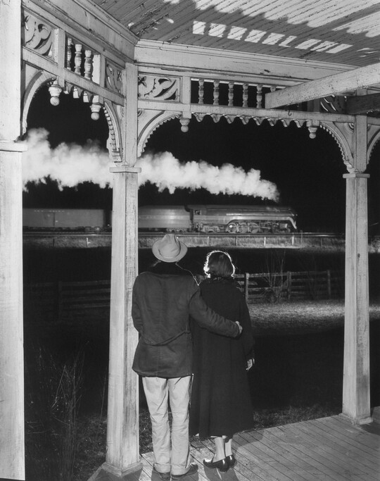 A black-and-white photograph of a couple standing under a covered porch watching a steam train pass by at night.