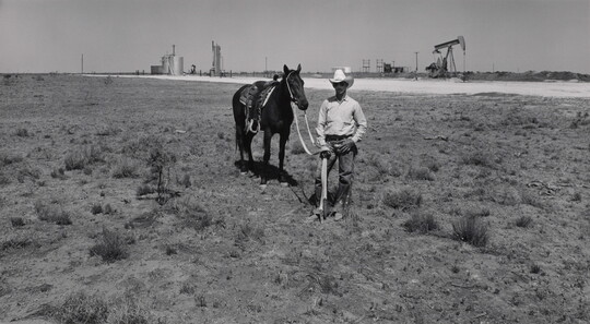 A black-and-white photograph of a man wearing sunglasses and a cowboy hat standing next to his horse, an oilfield and pumpjacks in the background.