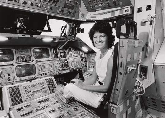 A black-and-white photograph of a White woman with short dark hair sitting in the cockpit of a space shuttle.
