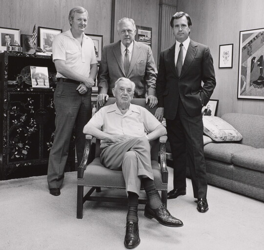 A black-and-white photograph of four White men of varying ages, three standing and one seated in an armchair, in an office.