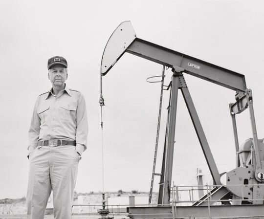 A black-and-white photograph of an older White man standing in front of a pump jack.