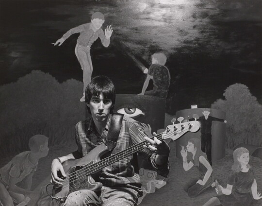A black-and-white photograph of a young White man playing an electric bass guitar in front of a mural of people smoking and drinking.
