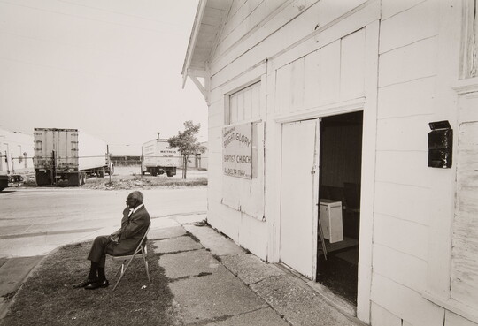 A black-and-white photograph of an older Black man in a suit sitting on a folding chair in front of a white building.