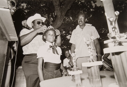 A black-and-white photograph of a young Black girl having a tiara placed on her head as others watch.