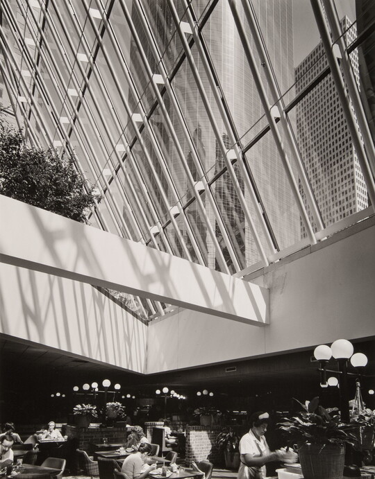 A black-and-white photograph of a cafeteria under a ceiling of windows that look up to a city skyline.
