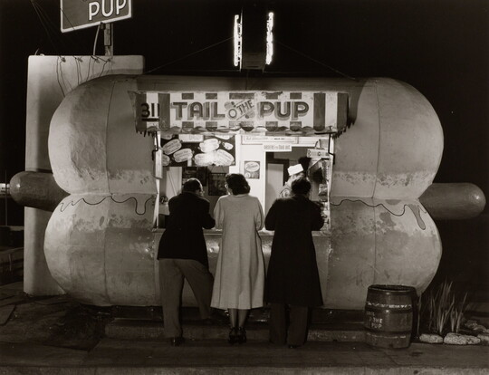 A black-and-white photograph of people ordering food at night at a hot dog stand shaped like a hot dog.