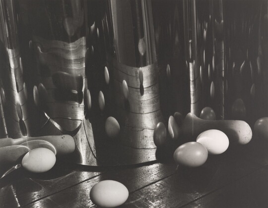 A black-and-white photograph of eggs on a wood surface reflected and distorted by an upright, wavy mirrored surface.