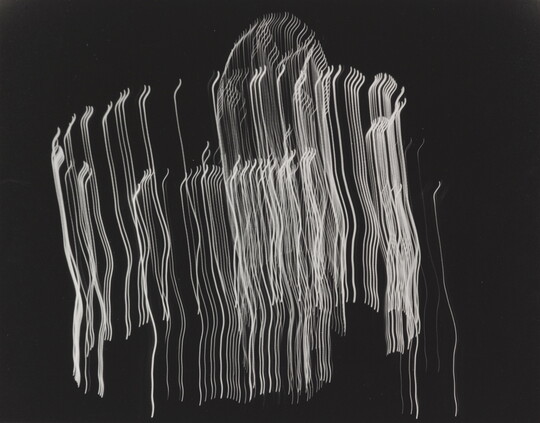 A black-and-white photograph of vertical streaks of white light layered in a circular shape on a black background.