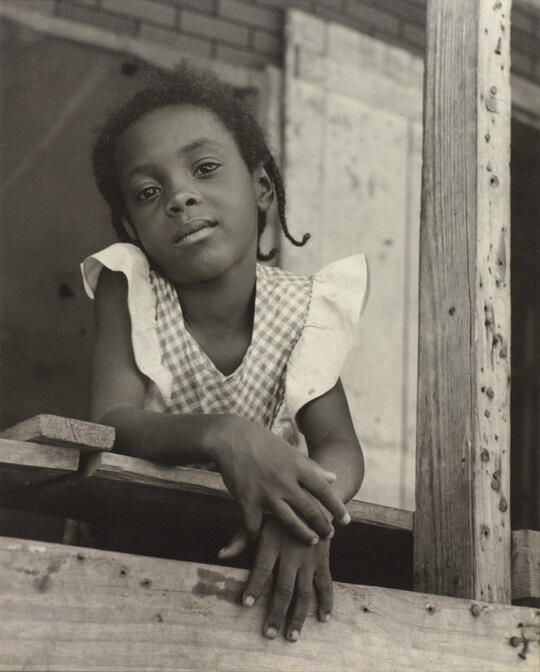 A black-and-white photograph of a young Black girl in a gingham top leaning out of a window.
