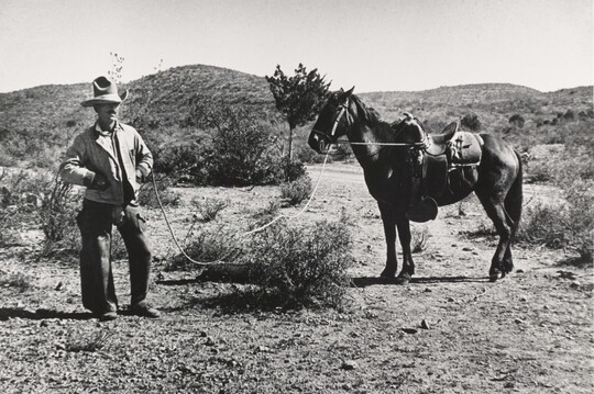 A black-and-white photograph of a White man wearing a large cowboy hat standing in the desert holding the reins of a saddled horse.