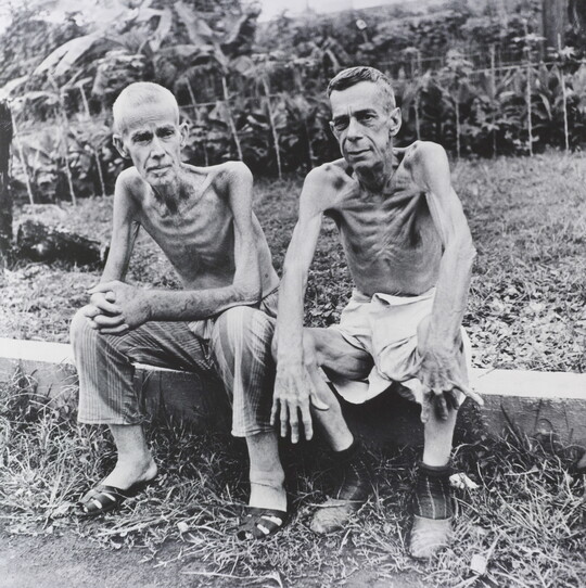 A black-and-white photograph of two shirtless, emaciated White men sitting on a concrete block.