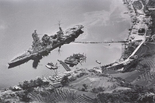 A black-and-white photograph of an aerial view of a large, damaged ship just off a hilly coast.