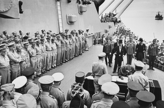 A black-and-white photograph of soldiers standing on the deck of a ship watching a smaller group of figures sign papers at a table.