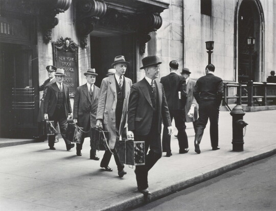 A black-and-white photograph of four White men in suits and hats exiting a city building and each holding a briefcase tethered to the other briefcases.
