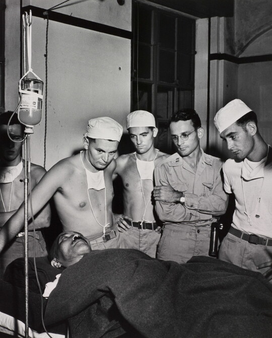 A black-and-white photograph of a man on a hospital bed hooked to an IV and under a blanket surrounded by five White men, some in military clothing, all watching him.