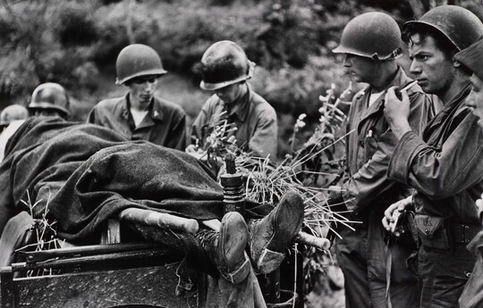 A black-and-white photograph of several helmeted White soldiers standing next a body covered in a blanket on a stretcher.