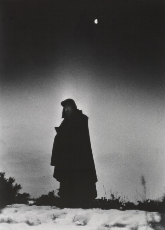 A black-and-white photograph of a heavily cloaked person standing in a frozen landscape with a half-moon behind them.