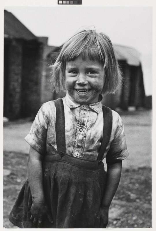 A black-and-white photograph of a smiling, coal-smudged young girl with fair skin and short hair, wearing in a dirty button-down shirt and skirt.