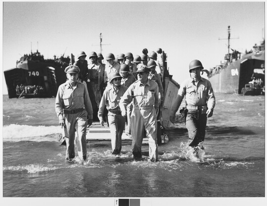 A black-and-white photograph of a large group of men in military uniforms, most wearing helmets, exiting boats and wading onto a shoreline.