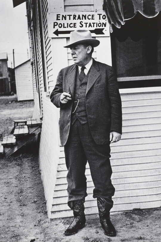 A black-and-white photograph of a White man in a three-piece suit, bowler hat, and cowboy boots, smoking a cigar and standing next to a clapboard building.
