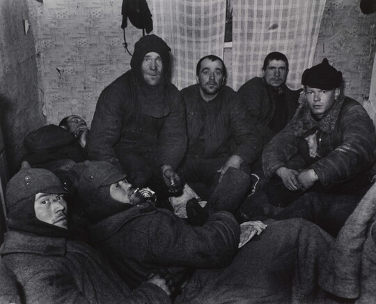 A black-and-white photograph of a group of White men wearing dark, heavy clothing huddled in a small room with plaid curtains over a window.