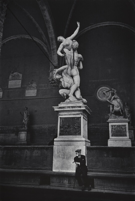 A black-and-white photograph of a man in a suit sitting below the statue of The Rape of a Sabine Woman in Florence, Italy.