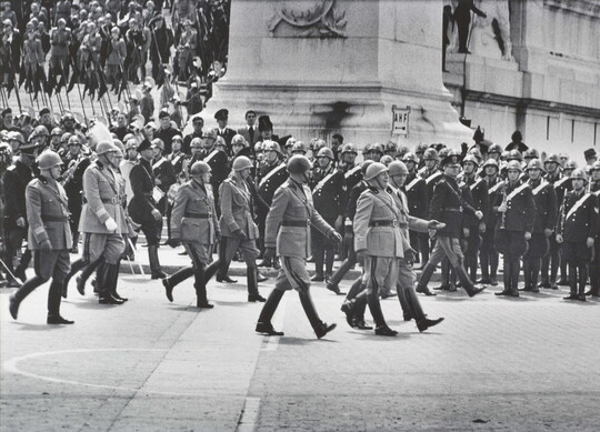 A black-and-white photograph of a military parade, with soldiers lined up to the side watching.
