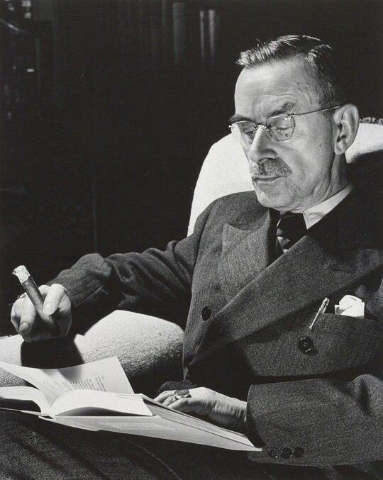 A black-and-white portrait photograph of a seated White man wearing a dark suit and glasses, holding a cigar, and reading a book.