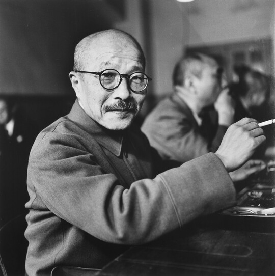 A black-and-white photograph of an Asian man with a shaved head, moustache, and round glasses holding a cigarette.
