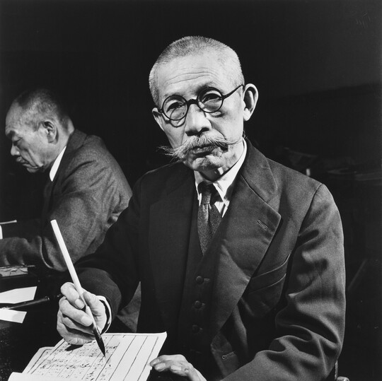 A black-and-white photograph of an Asian man with a shaved head, handlebar moustache, and round glasses writing on paper with a brush and ink.