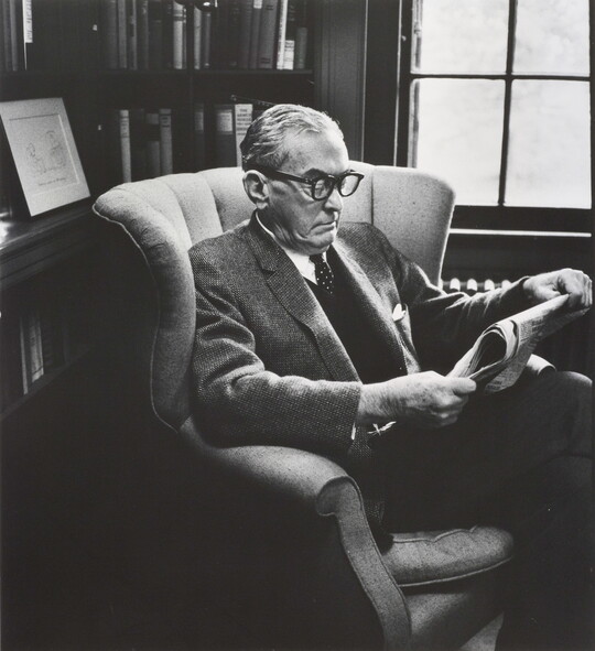 A black-and-white photograph of an older White man in business attire wearing thick, horn-rimmed glasses reading a newspaper in a wingback chair.