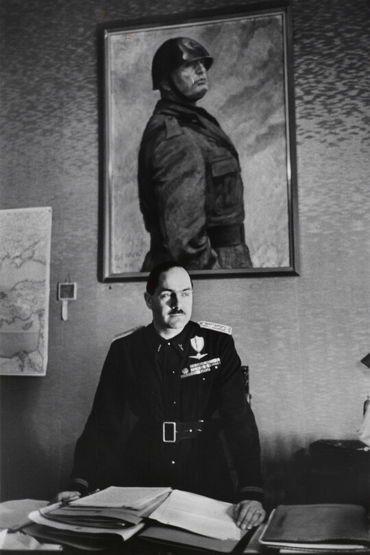 A black-and-white photograph of a White man in military regalia behind a desk stacked with papers above which hangs a portrait of a helmeted man in military dress.