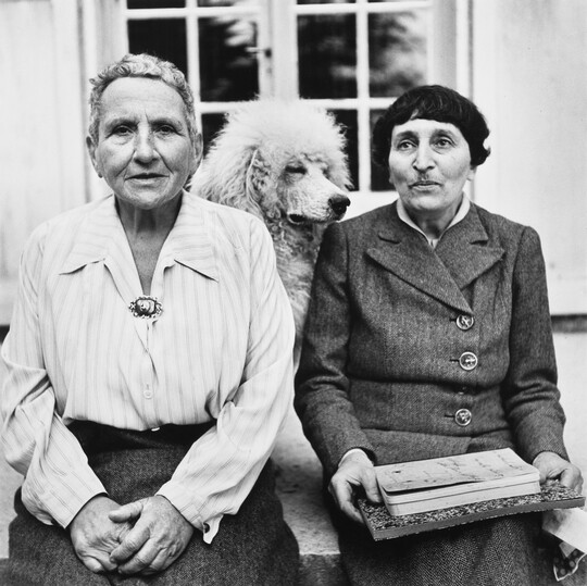 A black-and-white portrait photograph of two older White women seated with a dog in between them.