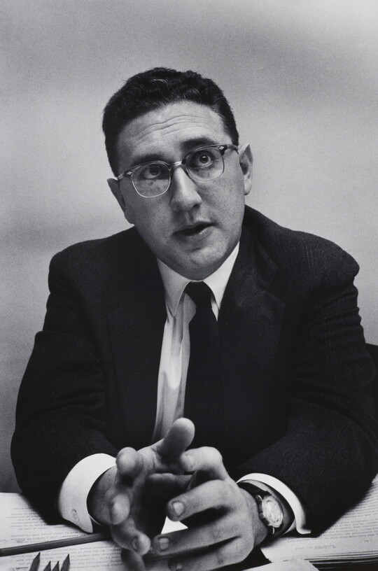 A black-and-white photograph of a White man with short, dark hair in a suit, tie, and glasses leaning forward with his elbows on a desk, gesturing with his hands.