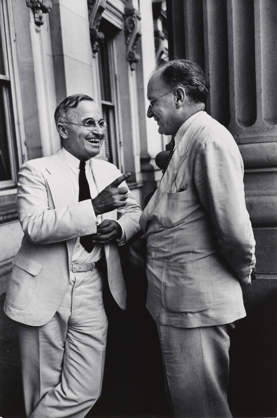 A black-and-white photograph of two White men, both in light-colored suits and glasses, talking to one another and smiling.