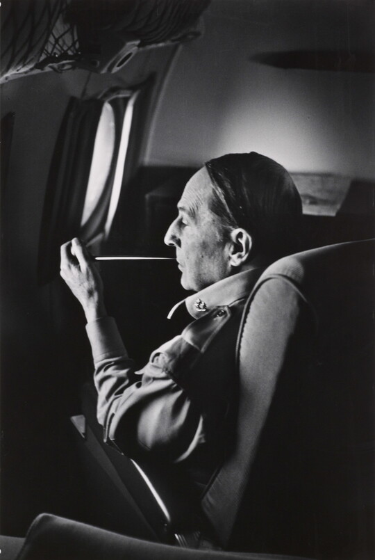 A black-and-white photograph of a White man seated on an airplane smoking a long thin pipe.