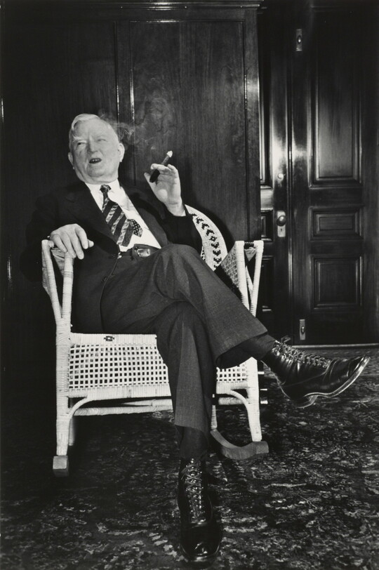 A black-and-white photograph of an older White man wearing a suit and smoking a cigar in a wicker rocking chair.