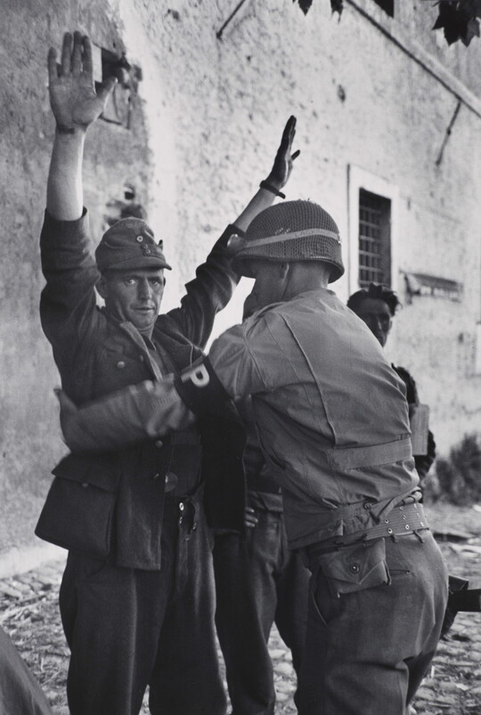 A black-and-white photograph of a White man in military uniform being frisked by a second man in military uniform.