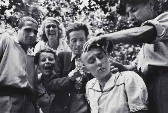 A black-and-white photograph of a seated White woman having her head shaved as laughing and jeering people watch.