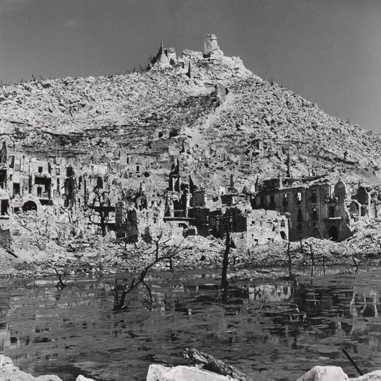 A black-and-white photograph of the ruins of a town at the edge of a body of water and a ruined building on top of a mountain in the background.
